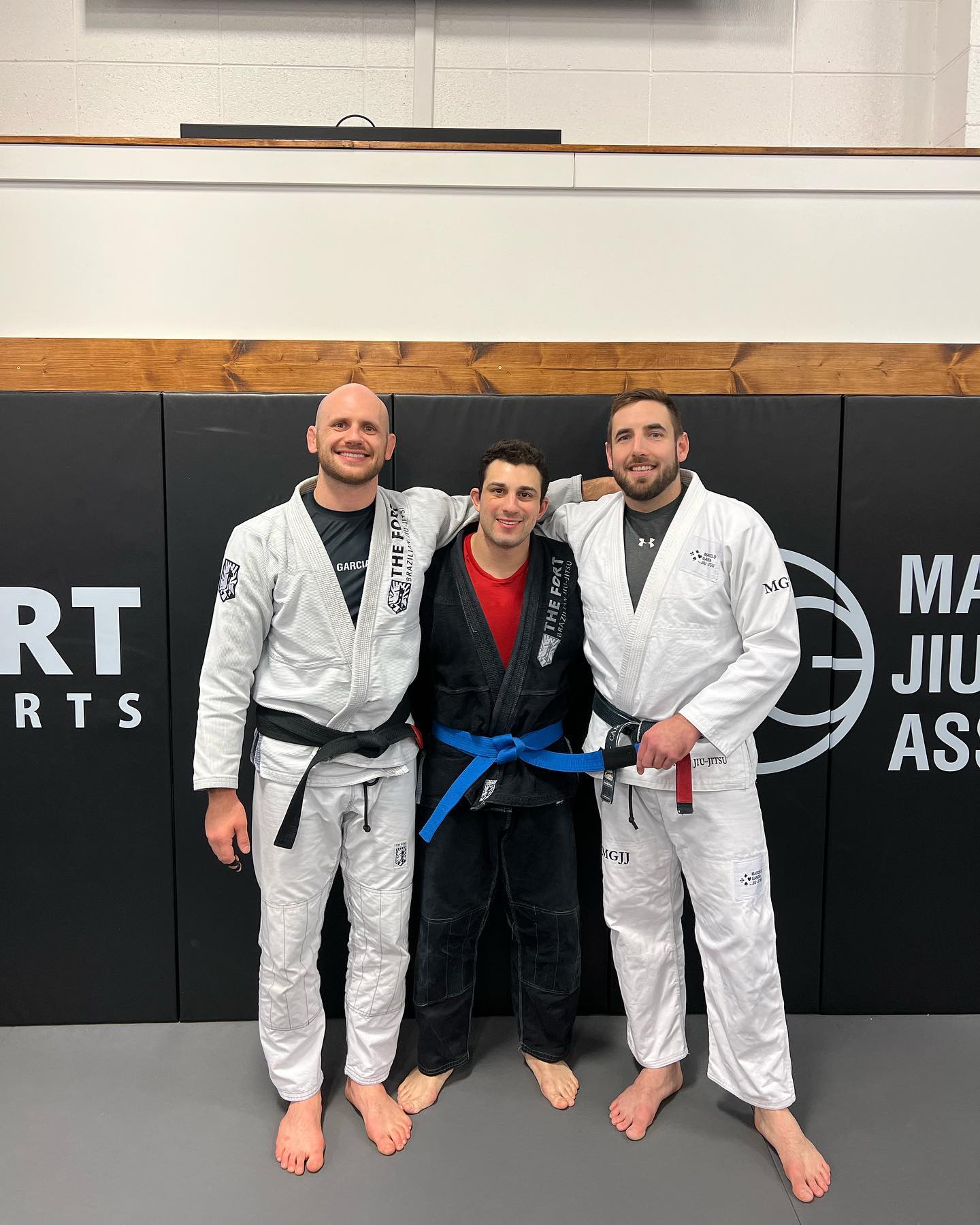 A member wearing a blue belt stands between two black-belt coaches in front of The Fort wall.