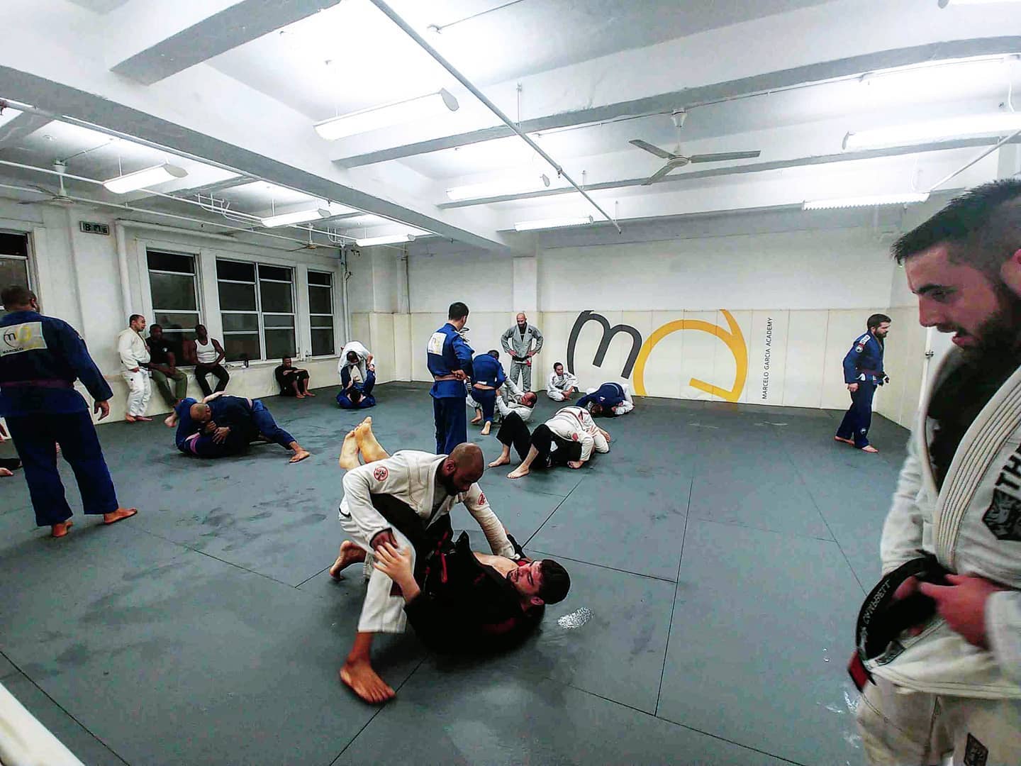 Several pairs of grapplers train in front of the Marcelo Garcia Academy wall logo, with a Fort-branded gi visible in the foreground.