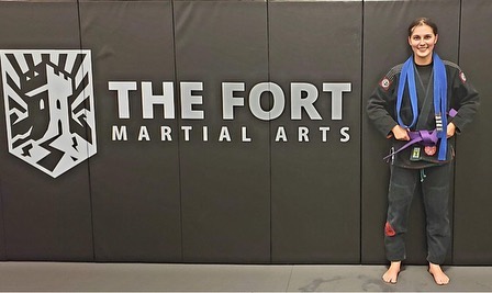 A member wearing a purple belt poses beside The Fort Martial Arts wall logo.